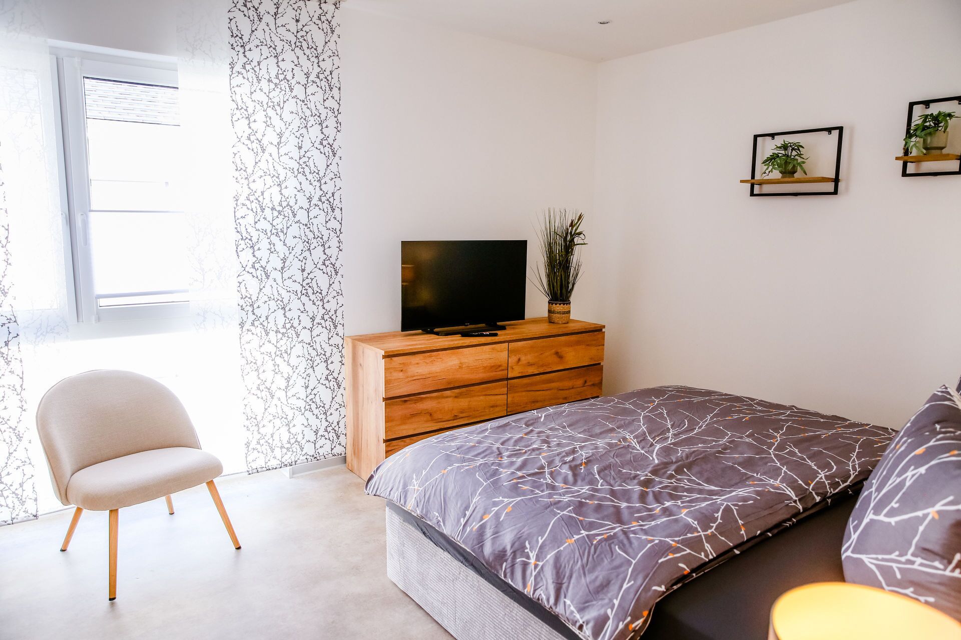 Un dormitorio con una cama, un televisor sobre un aparador de madera, una silla y dos plantas en estanterías de pared.