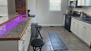 A modern kitchen with a central island, bar stools, a ceiling fan, and a brick accent wall. - Cozy Historic Home just 2 miles from USC, downtown and 6 miles from Ft. Jackson (Columbia)