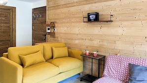 Uma sala de estar com um sofá amarelo, uma poltrona estampada, uma mesinha com xícaras e uma televisão em uma parede de madeira.