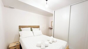 Cama bem arrumada com lençóis e toalhas brancas, mesa de cabeceira de madeira e guarda-roupa branco.
