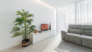 A modern living room with a grey sofa, a large potted plant, a flat-screen TV on a white console, and sheer curtains on the window. - GuestReady - Urban chic in Aveiro (Aveiro)