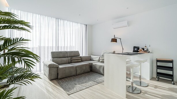 A modern living room with a grey sectional sofa, a small white bar area, and a large window with sheer curtains. - GuestReady - Urban chic in Aveiro (Aveiro)