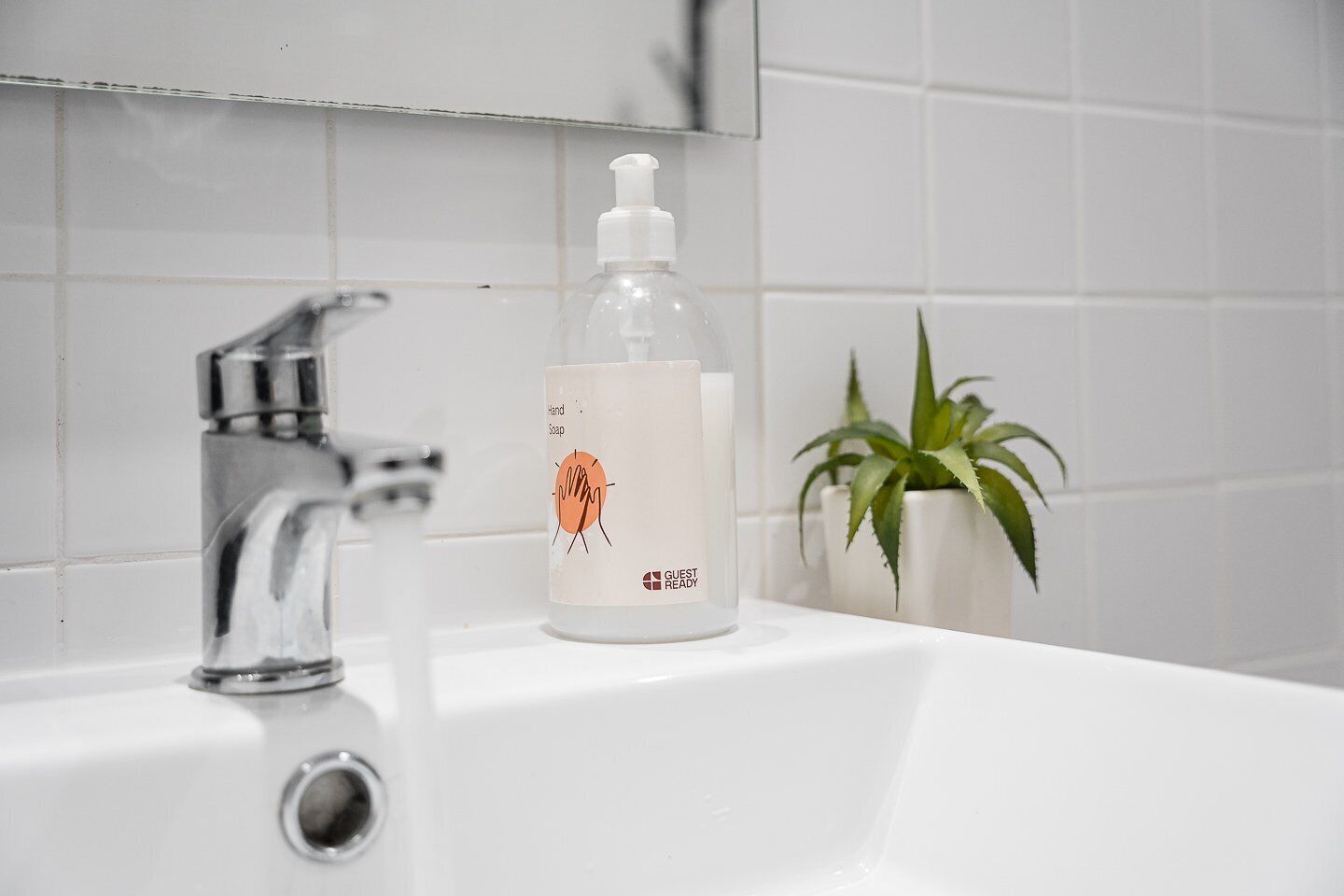 A bathroom sink with a clear glass bottle of hand soap labeled "Guest Ready" and a potted plant.