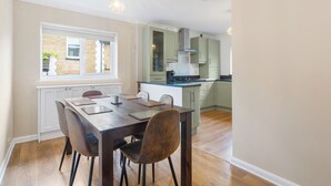 A modern kitchen with white cabinets, a wooden dining table with chairs, and a window with a view of a brick building. - Modern Serenity in Langley Vale (Epsom)