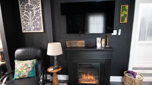Een zwarte leren fauteuil met een gebloemd kussen, een staande lamp, een open haard met vuur, een flatscreen-televisie en lijstwerken aan de muur.