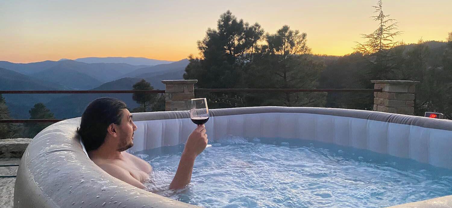 A person in a hot tub with a glass of red wine, surrounded by a mountainous landscape.