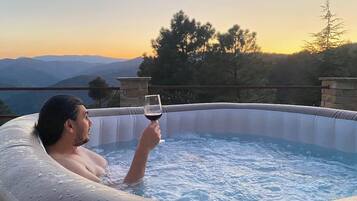 A person in a hot tub with a glass of red wine, surrounded by a mountainous landscape.