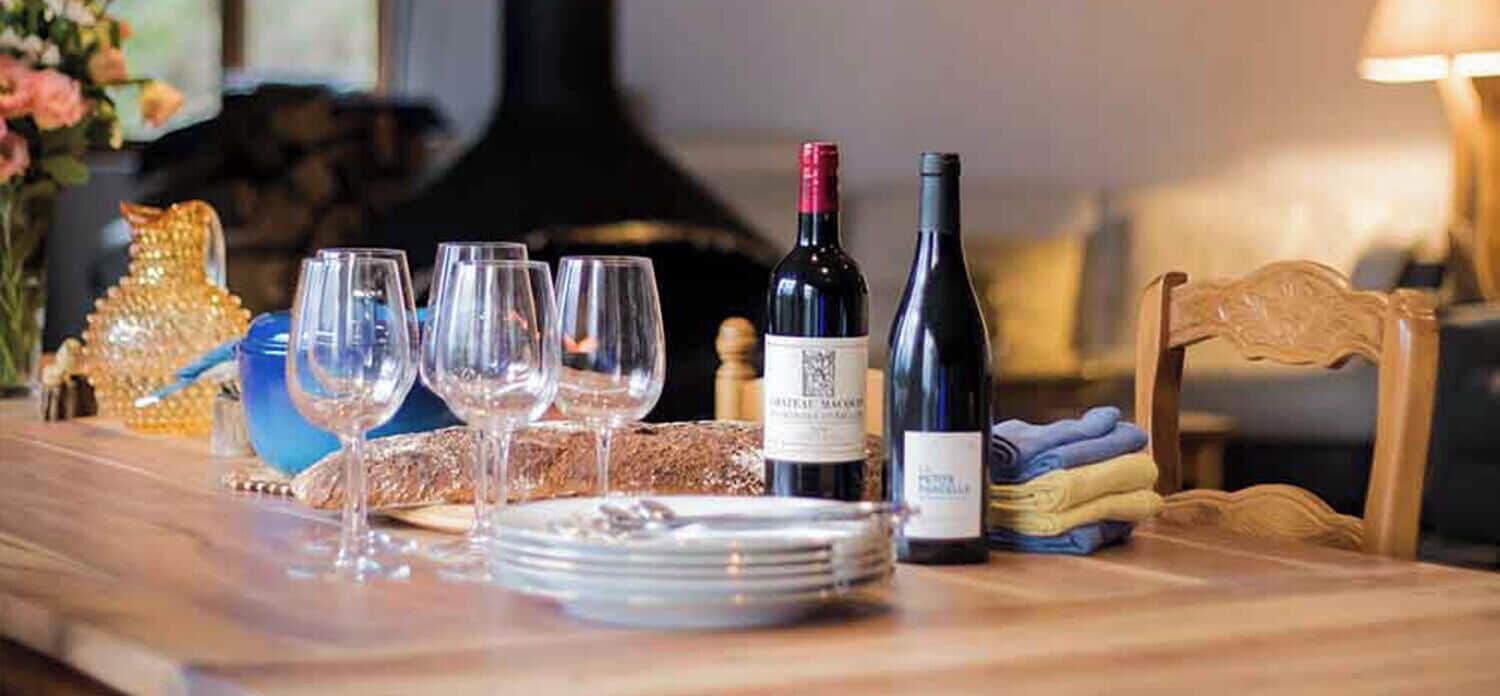 Una mesa de comedor con botellas de vino, copas y platos, en una habitación con chimenea y mobiliario de madera.