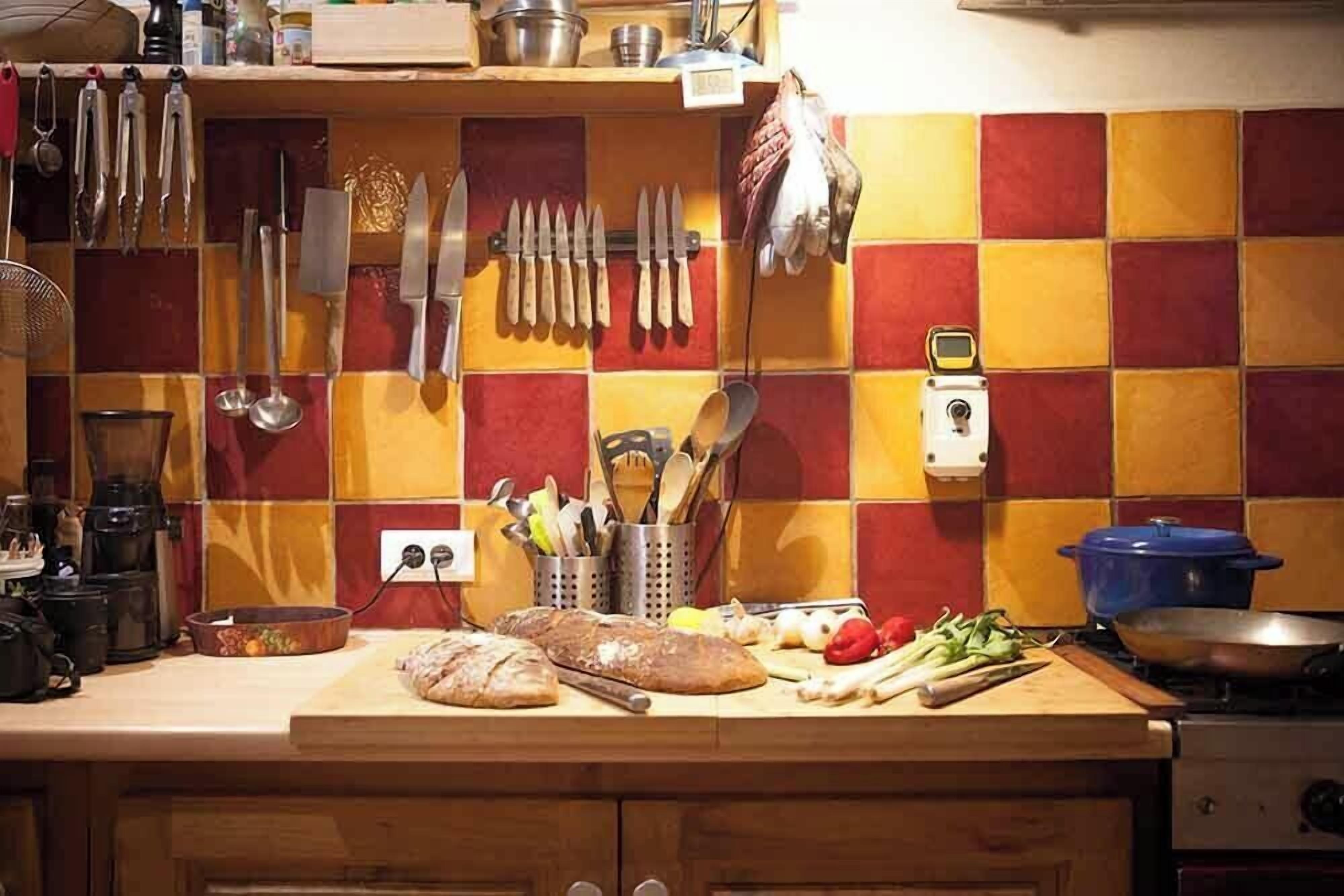 Una cocina con un salpicadero de baldosas a cuadros, varios utensilios colgando y una encimera con pan, verduras y una olla azul.