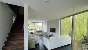 Ein modernes Wohnzimmer mit Treppe, weißen Sofas, einem Flachbildfernseher und großen Fenstern.