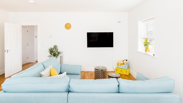 Een moderne woonkamer met een lichtblauwe modulaire bank, een flatscreen televisie aan de muur en een klein bijzettafeltje met decoratieve objecten.