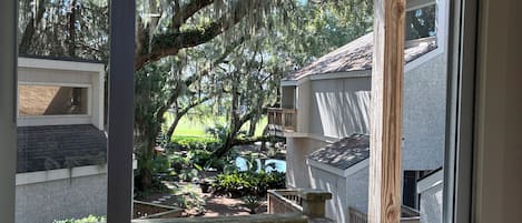 A view from a balcony with a wooden railing, a glass door, and a view of a garden with trees and a building in the background.