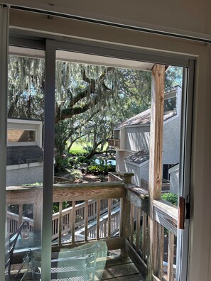 A view from a balcony with a wooden railing, a glass door, and a view of a garden with trees and a building in the background. - Cozy 2-bedroom condo with sunroom on a beautiful golf course St. Simons Island.
 (St. Simons Island)