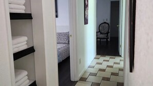 Un pasillo con suelo de baldosas, una cama con colcha estampada y una silla al fondo.