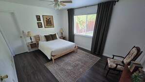 Un dormitorio con cama, una silla, escritorio, ventana con cortinas, ventilador de techo y cuadros en la pared.
