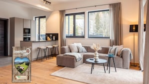 A modern living room with a sofa, ottoman, and coffee table, a kitchen area with bar stools, and large windows offering a view of trees. - Penthouse With Shared Pool (Sankt Martin am Tennengebirge)