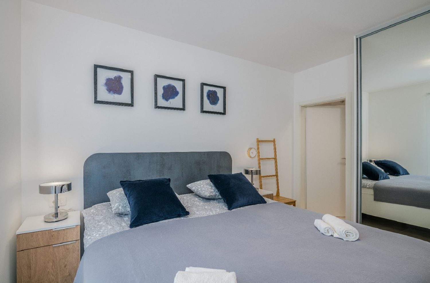 A modern bedroom with a large bed, a bedside table, a lamp, and a mirror.