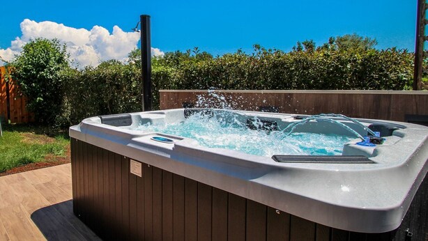 Een jacuzzi met waterstralen, omringd door een houten terras en een houten omheining, met een helderblauwe lucht en bomen op de achtergrond.