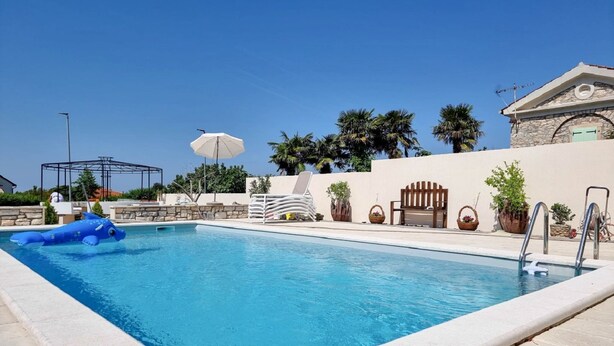 Une piscine entourée d’un mur de pierre et de palmiers, avec des chaises longues et des parasols.