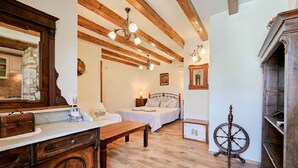 Une chambre avec des poutres en bois, un lit avec une tête de lit, une table de chevet, une commode, un miroir, un lustre et un rouet.