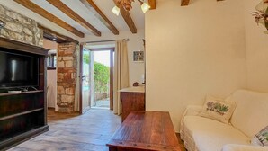 Un salon confortable avec une télévision, un banc en bois et un canapé. On y trouve un pilier en pierre et une porte qui donne sur l’extérieur.