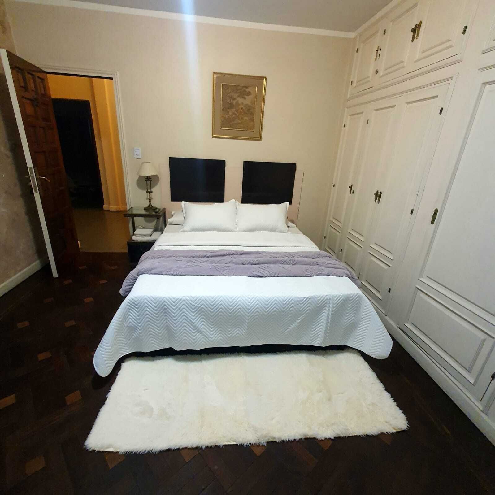 Una habitación de hotel con cama, mesita de noche, lámpara, alfombra blanca y un armario.