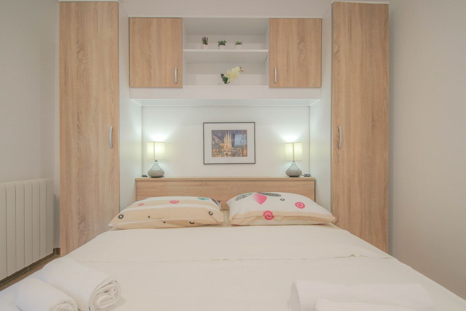 Une chambre à coucher comprenant un lit, deux tables de chevet, une tête de lit, un tableau encadré et un radiateur.
