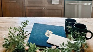 A kitchen with a window, wooden cabinets, a sink, and a table with a blue notebook, a white envelope, and two black mugs.