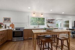 Una cocina con gabinetes de madera, un horno de acero inoxidable, una isla grande con taburetes y un ventanal con vista a la vegetación.