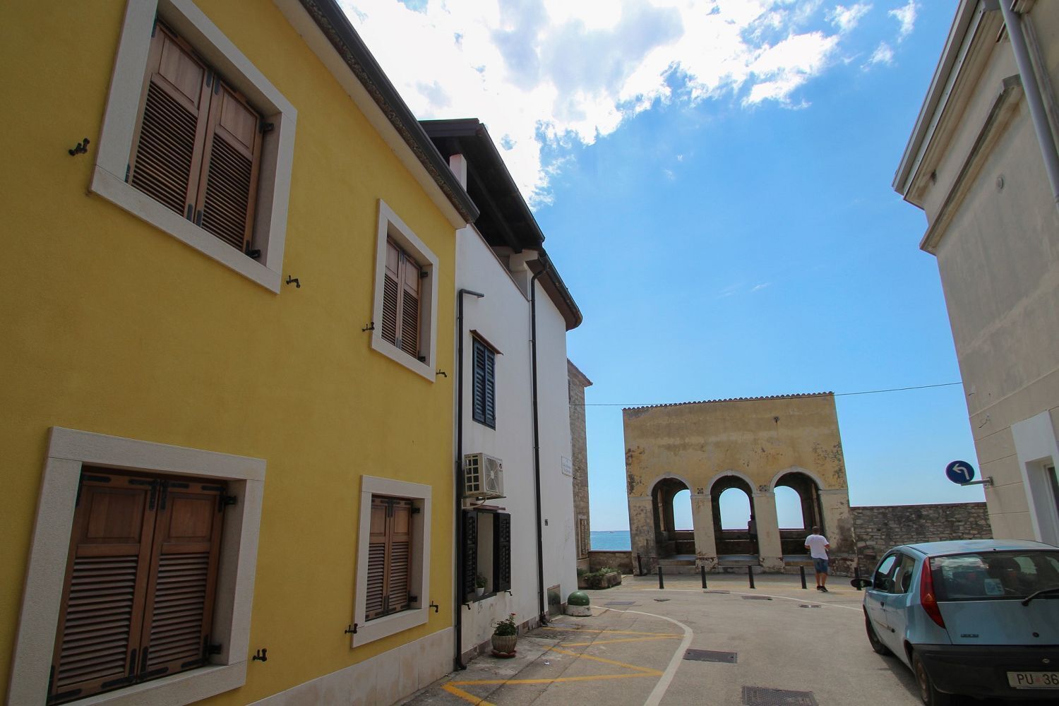 A narrow street with yellow and white buildings, a blue car parked on the right, and a historical stone structure with arches in the background.