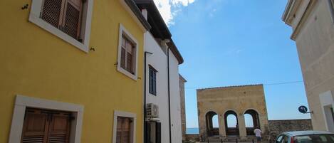 A narrow street with yellow and white buildings, a blue car parked on the right, and a historical stone structure with arches in the background.