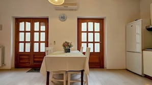 Un comedor con una mesa dispuesta para dos personas, dos puertas de madera con paneles de vidrio esmerilado, un reloj de pared y un refrigerador blanco.