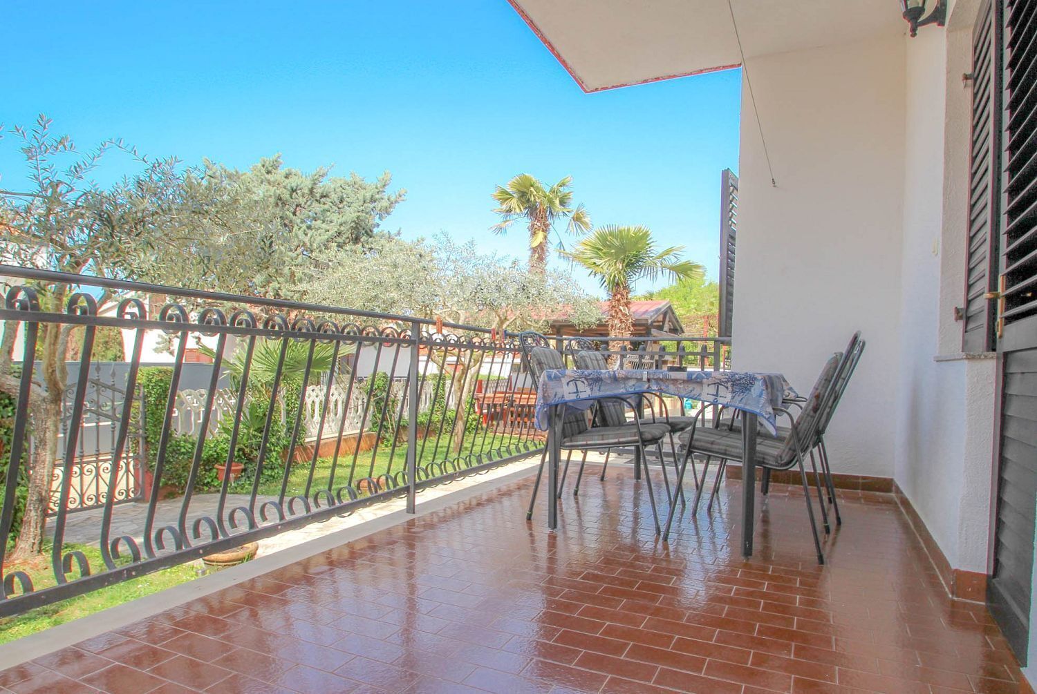 A covered patio with a table and chairs, a metal fence, and a view of a garden with trees and a palm tree.