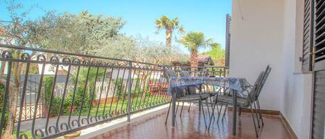 A covered patio with a table and chairs, a metal fence, and a view of a garden with trees and a palm tree.
