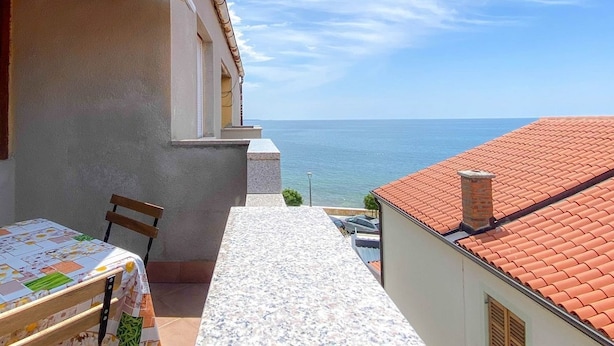 A narrow outdoor corridor with a table and chairs, leading to a balcony overlooking the sea.