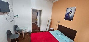 A hotel room with a bed, a TV, a desk with chairs, and a door leading to a hallway. - MAGIC HOSTAL (Quito)