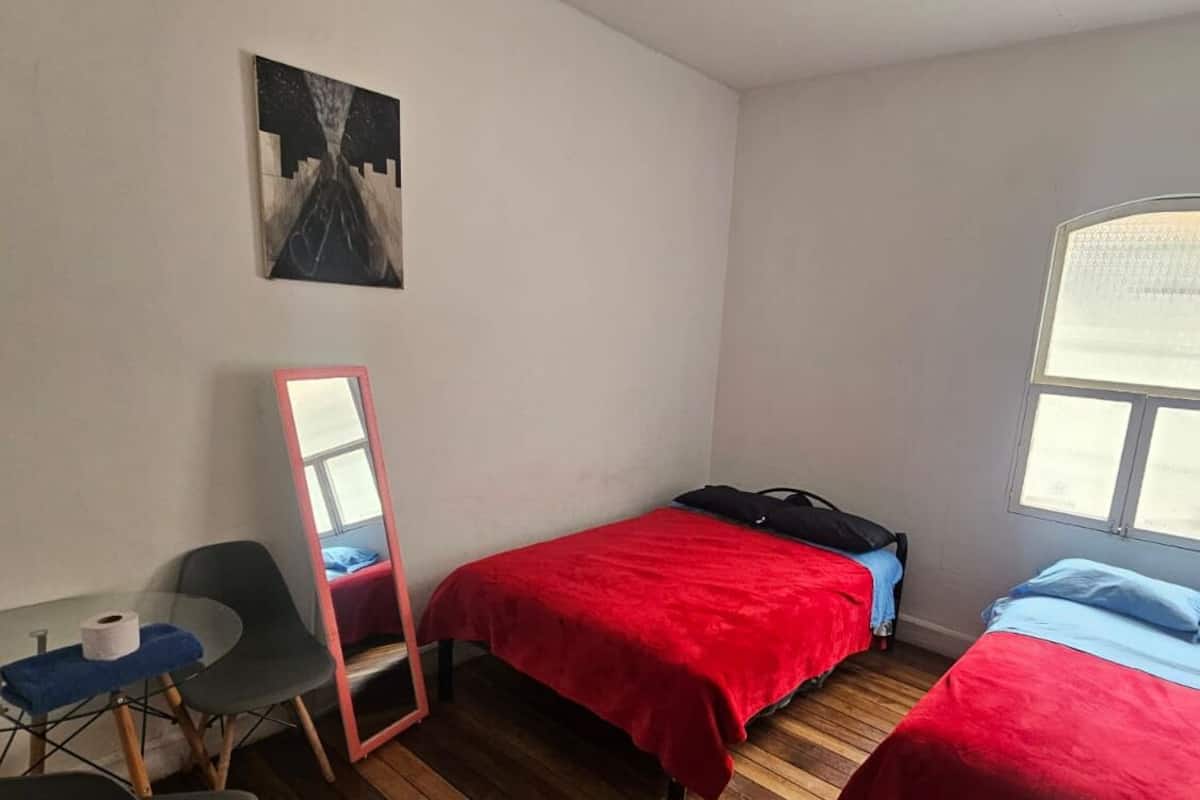 A small, sparsely furnished room with two beds, a desk, a chair, a mirror, and a window.