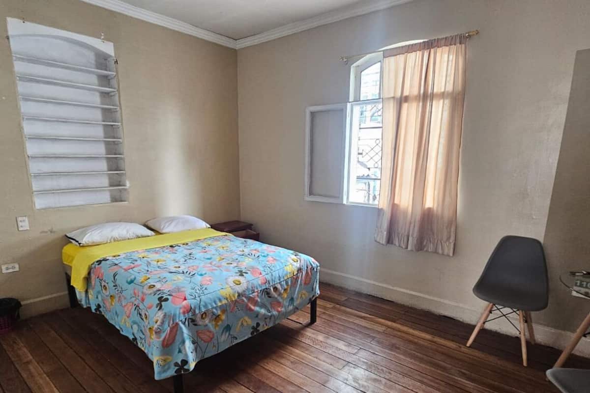 A bedroom with a bed, a window with curtains, a chair, and a small table.