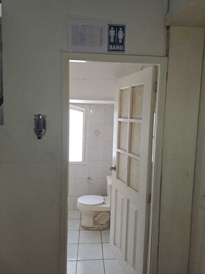A bathroom with a toilet, a sink, and a shower. There is a sign indicating a men's and women's restroom. - MAGIC HOSTAL (Quito)