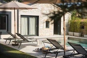 Un espace aménagé au bord de la piscine, avec des chaises longues, une table où sont posées des boissons, et un bâtiment en pierre doté de grandes fenêtres.