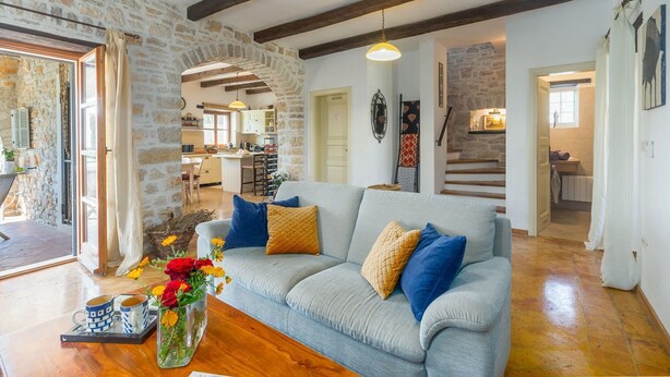 Un salon chaleureux avec un canapé bleu clair, des poutres en bois, des murs en pierre, une cuisine à l’arrière-plan et un escalier menant à un autre niveau.