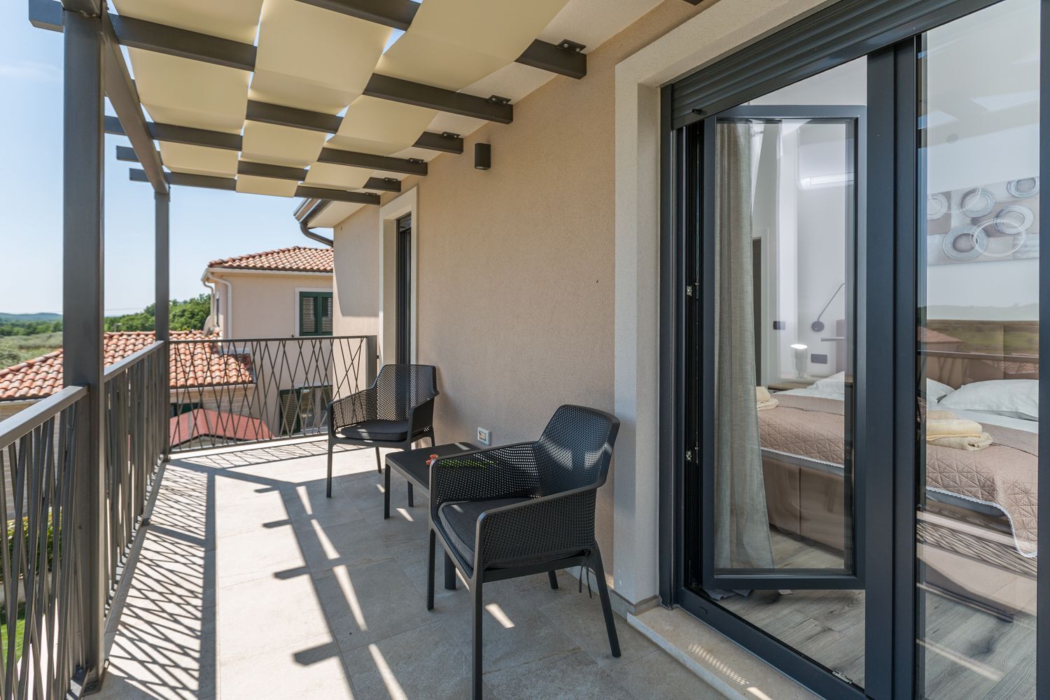 A balcony with two chairs and a table, a sliding glass door leading to a bedroom with a bed and a nightstand.