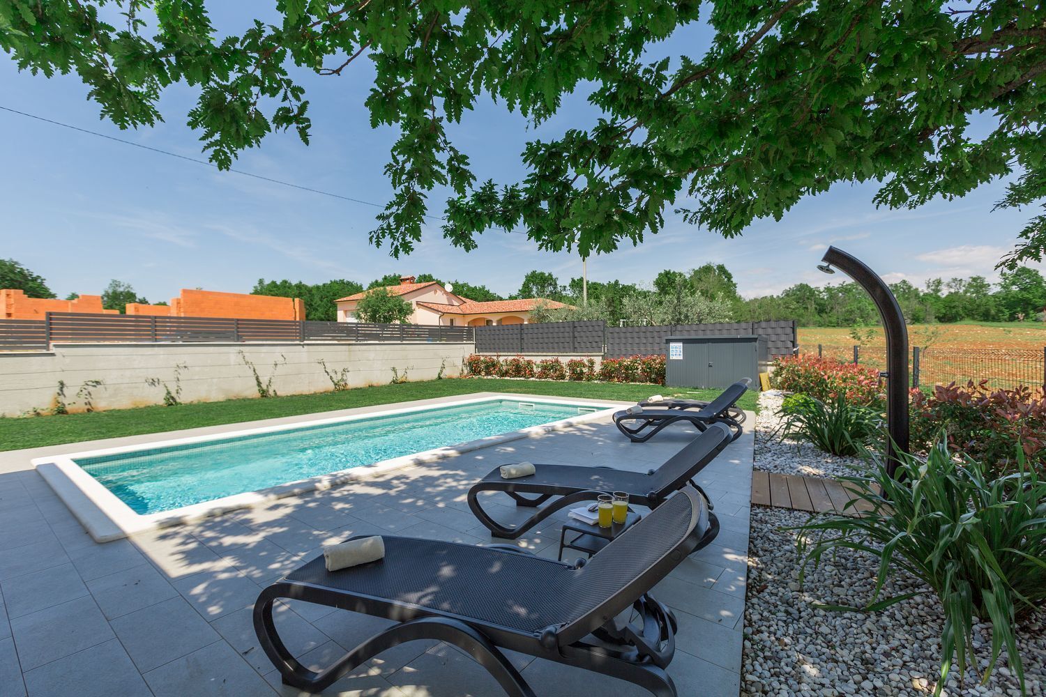 A swimming pool with two lounge chairs and a small table with drinks, surrounded by a garden and a fence.
