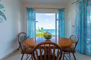 Un comedor con mesa y sillas de madera, un cuenco de fruta y vistas al océano a través de una ventana con cortinas florales.