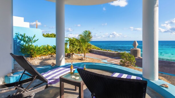 A covered outdoor seating area with a view of the ocean, featuring lounge chairs, a table, and a bottle of beer.