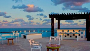 An outdoor seating area with a pergola, chairs, and a table, overlooking a beach and the sea.