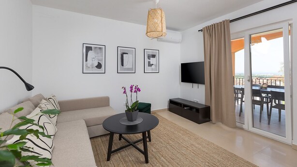 A modern living room with a sofa, a round coffee table with orchids, a TV, and a balcony with outdoor furniture.