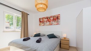 A modern bedroom with a large bed, a bedside table, a nightstand, a hanging light fixture, and a window with curtains.