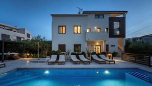 Une maison moderne à deux étages, dotée d’une piscine, de chaises longues et d’une entrée bien éclairée.
