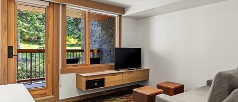 A modern living room with a stone wall, wooden paneling, a flat-screen TV, and a view of greenery through large windows.